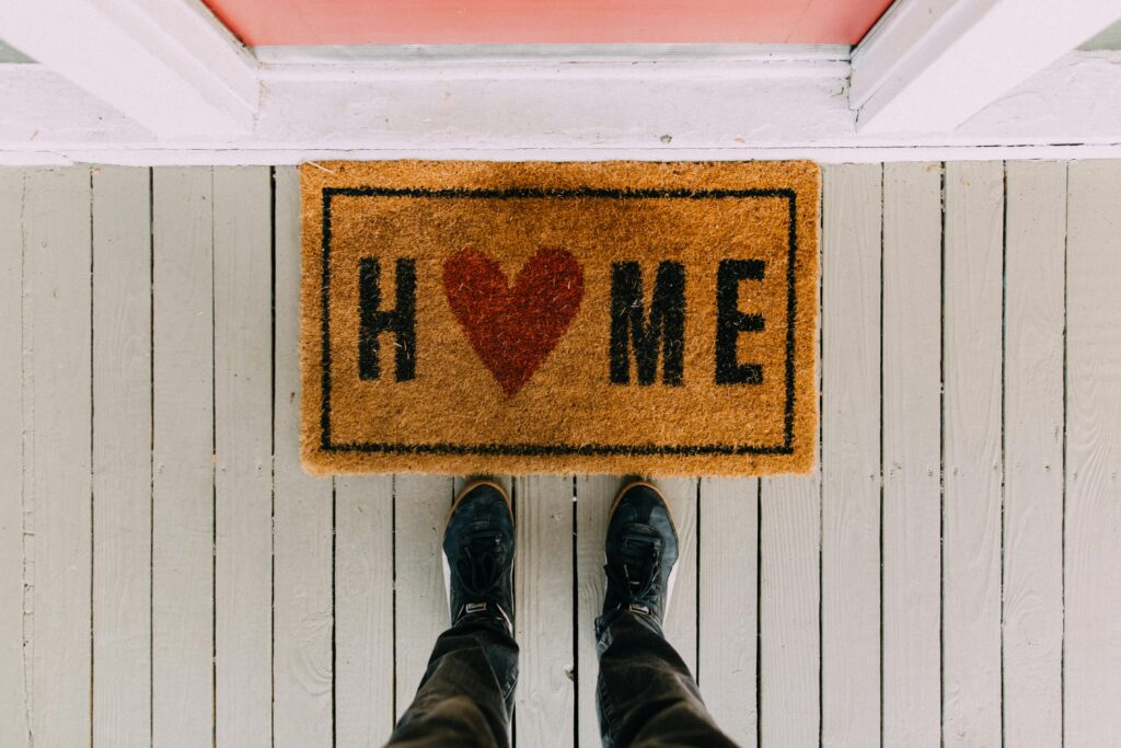 Close-up of doormat that says Home, with a heart for the O, and a pair of feet standing on its edge.