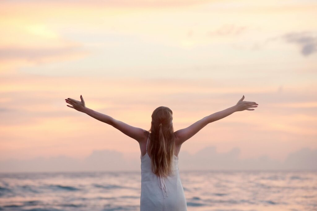 A person holding their arms out against a pink sky above the ocean.