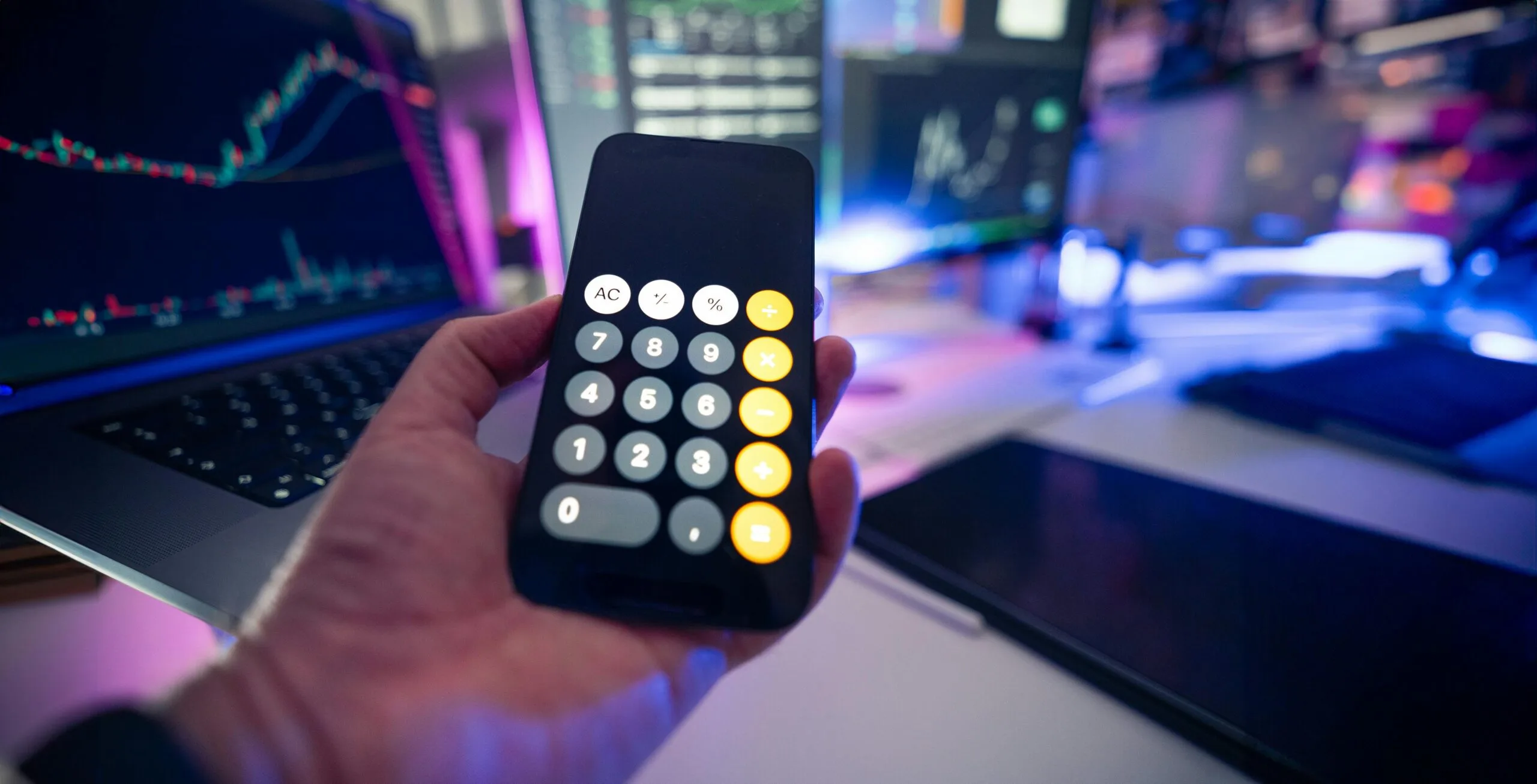 Closeup of a calculator glowing against a dark background showing a laptop screen with a financial chart.